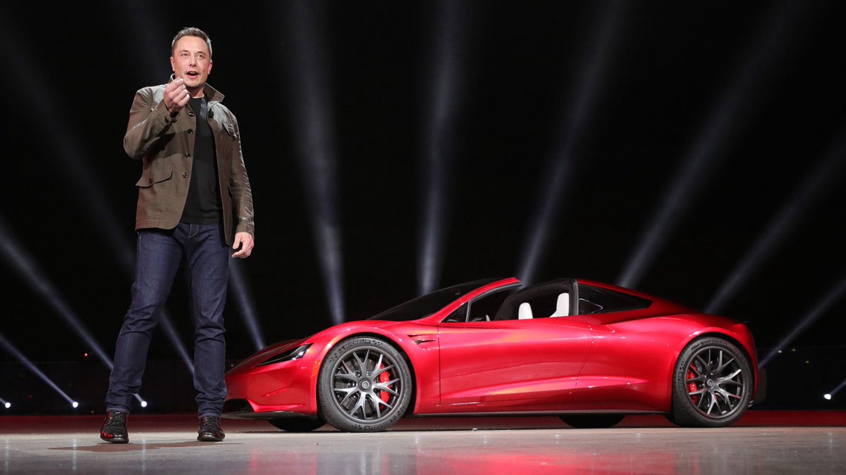 Elon Musk standing beside a red Tesla Roadster prototype at a reveal event; historical event photo, not a current trim-price chart